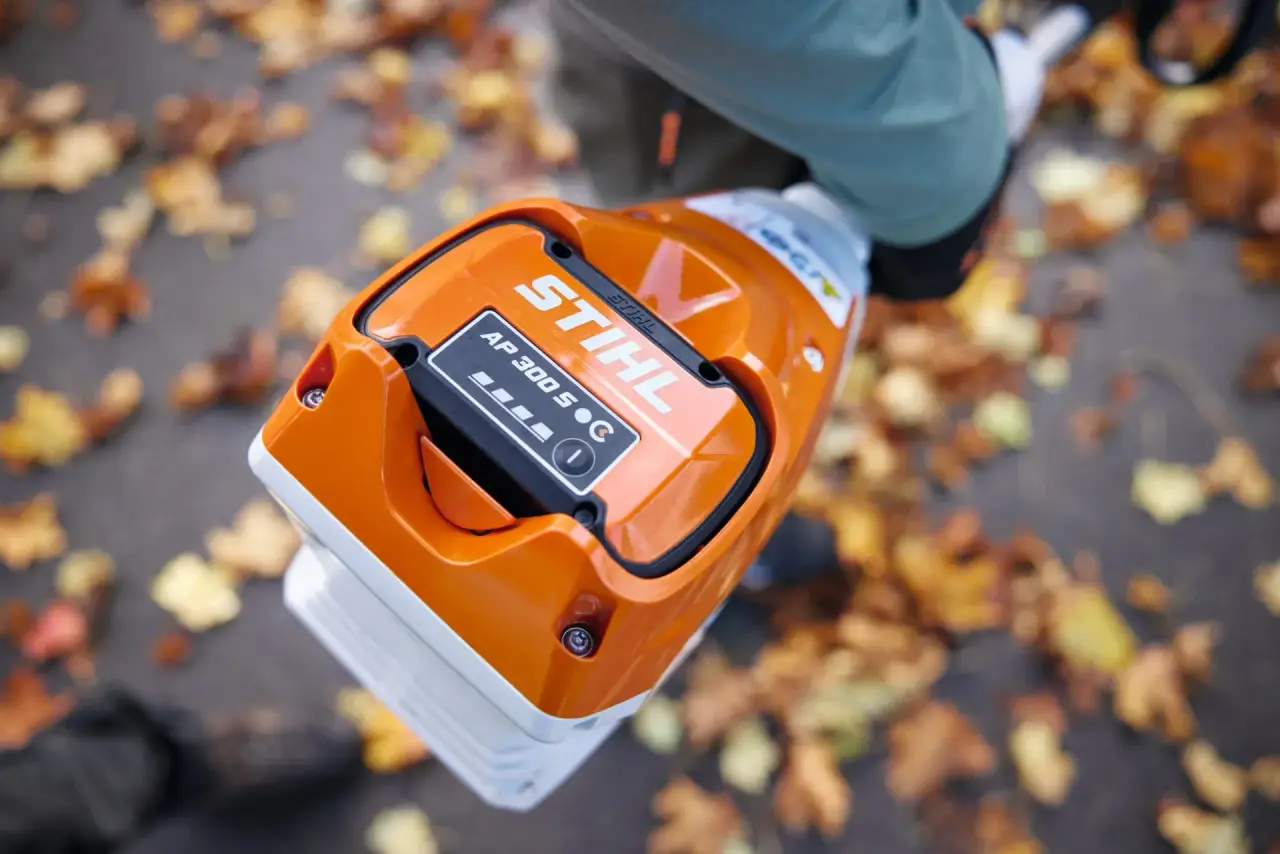 Close-up of a STIHL AP 300 S battery being carried outdoors over a path covered in fallen leaves